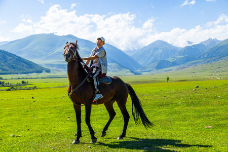 Semenovskoye Gorge, Kyrgyzstan - July 17 2024 - Young Asian man riding brown horse in sunny grassy fieldのeditorial素材