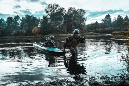 Covert landing. Pair of armed operators with painted faces disembarking river coast from military kayak . Diversionary mission under cover of darknessの写真素材