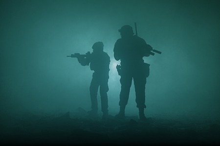 Black silhouettes of pair of soldiers in the smoke haze moving in battle operation. Back lightの写真素材