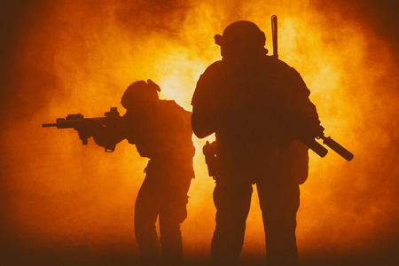 Black silhouettes of pair of soldiers in smoke moving through fire burning in battle operation, sacrificing their lives of our secutiry. Back lightの写真素材