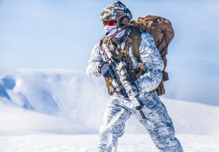 Army serviceman in winter camo somewhere in the Arctic. He wears chest rig, backpack, suffers from extreme cold, strong wind, but endures while mission continues, running moving across snow desertの写真素材
