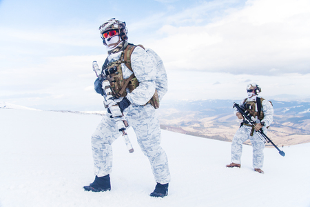 Army servicemen in winter camo somewhere in the mountains. Walking moving across snow desert despite bad weather wind and cold, holding weapons - assault and sniper rifle. Full body portraitの写真素材