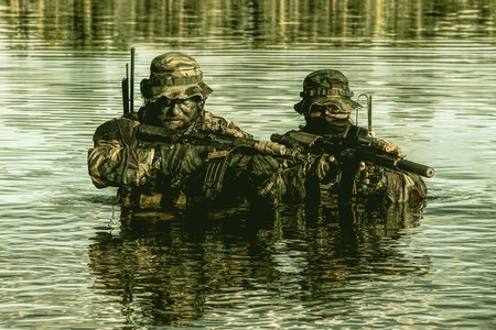 Pair of soldiers in action during river raid in the jungle waist deep in the water and mud and...