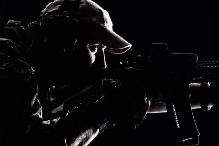 Studio contour backlight shot of special forces soldier in uniforms and baseball cap, pointing rifle, closeup portrait on black backgroundの写真素材