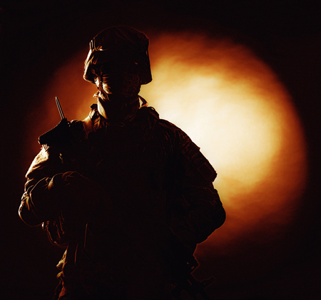 Silhouette photo of army soldier, modern combatant, military conflict participant standing in combat uniform with fiery spot on black background. Fire of war, ashes of battle, nuclear fire conceptの写真素材
