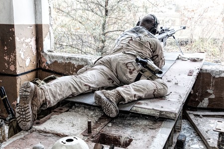 Portrait of U.S. Navy SEAL sniper on firing position, armed with large caliber sniper rifle with telescopic sight, wearing tactical headset with microphone, observing territory, searching targetsの写真素材