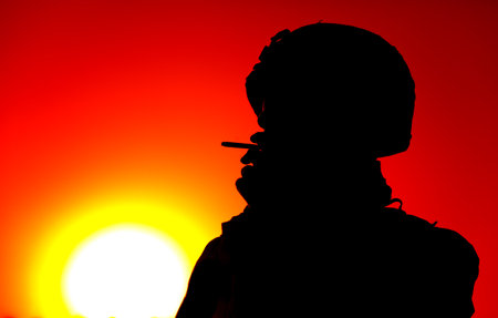 Silhouette of army soldier in combat helmet smoking cigarette on background of red sunset sky and...