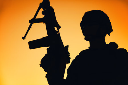 Shoulder silhouette of army machine gunner, special operations forces infantryman standing with raised gun on background of sunset sky. Marine Corps shooter in combat helmet holding weapon at dawnの写真素材