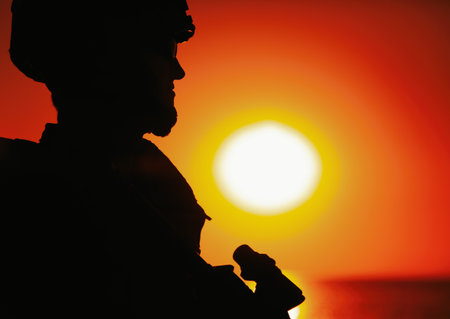 Shoulder silhouette of army soldier, special operations forces fighter in combat helmet and ballistic glasses, standing with gun on background of setting below ocean horizon sunの写真素材