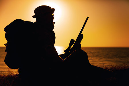 Silhouette of commando fighter, army special forces sniper sitting on sea or ocean shore on sunset. Coast guard rifleman observing beach, resting during coastline patrol or amphibious operationの写真素材