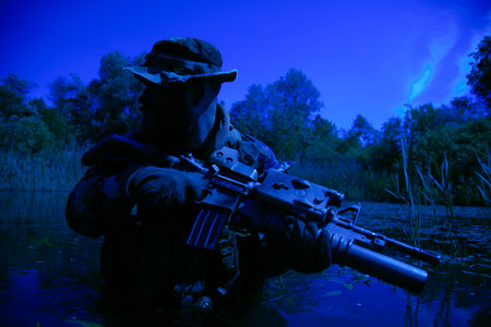 A focused soldier surveys the marshland with his rifle, submerging himself, dark night, his arms and...