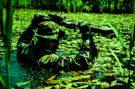 A camouflaged soldier walks through a swamp, submerging himself, his arms and rifle visible, shot from behind, realities of harsh military serviceの写真素材