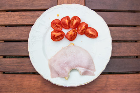 Snack on a white plate on a wooden table. Red tomatoes and bitten toat with ham and butter.の写真素材