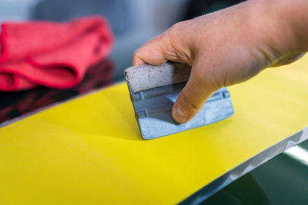 Close-up of a person's hand installing a vinyl wrap on a car. Tools for installing designer wraps made of yellow self-adhesive wrap.の写真素材