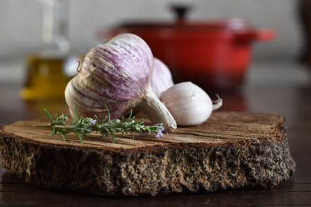 organic ingredients: fresh garlic, rosemary branch on rustic wood. in the background, out of focus, olive oil bottle and red pan. Selective focus.の写真素材