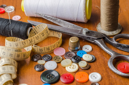 A collection of sewing tools and supplies in a sewing kit, on woodem table. Items include threads, buttons, needle, tape measure, scissors, and thimble.の写真素材