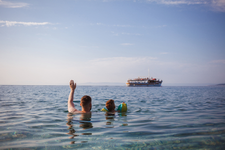 father and son waving their arms out of the water at a passing shipの写真素材