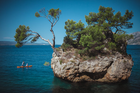 View from the shore to the Stone brela. A man and a girl are swimming on a board with a paddle around a rock.のeditorial素材