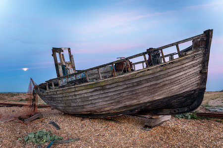 An old wrecked wooden fishing boat on a shingle beach under the light of a full moonの写真素材