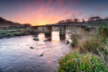 Dramatic vibrant sunrise over an ancient granite clapper bridge at Postbirdge on Dartmoor in Devon