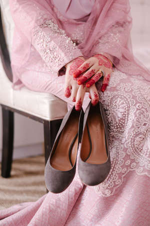 Kuala Lumpur, Malaysia - March 16, 2020: Malay Wedding, The bride sitting on a white chair with henna in hand holding beautiful of dark grey high heels, with light pink wedding dress floral patternのeditorial素材