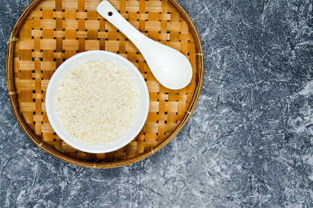 A long grain white rice, on rattan handmade bowl with wooden spoon isolated and black walnut chopping board on concrete pattern wall background. Copy spaceの写真素材