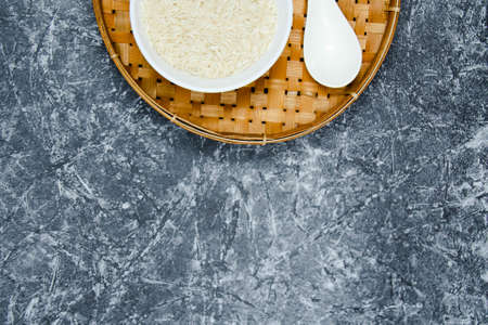 A long grain white rice, on rattan handmade bowl with wooden spoon isolated and black walnut chopping board on concrete pattern wall background. Copy spaceの写真素材