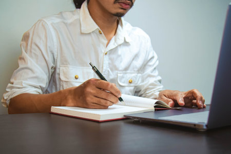 A young man with mustache wearing white shirt looking to the laptop and writing on white paper. Copy...