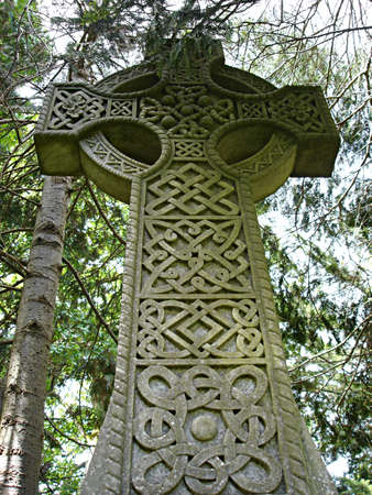 An old stone celtic cross, green with moss, rises up into its wooded surroundingsの写真素材
