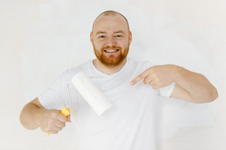 Young smiling employee handyman man wear t-shirt point index finger on paint roller.Repair home.の写真素材
