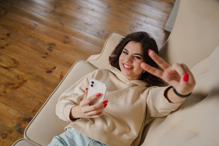 Cheerful young woman sincerely smiles and shows peace sign. Attractive brunette lady.の写真素材