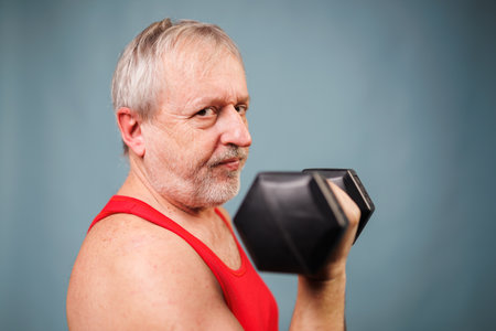 Senior Man Poses with Dumbbells in Blue Studioの写真素材