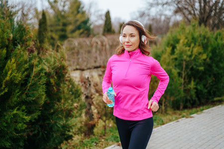 A hispanic runner jogging on a sunny day, pausing for a moment to drink water from her bottle.