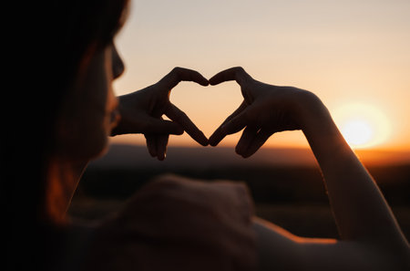 Hands shape heart. Little girl make sign love hands sunset. Small kid draw heart hands against sky. Sign heart symbol love, feeling. Silhouette of heart hands in sun close-up.Sunlight between fingersの写真素材