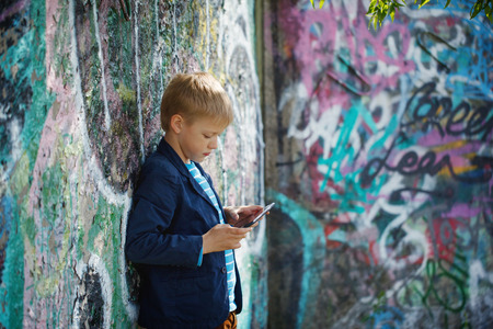 Little kid  absorbed into his tablet for educating and playing.Boy standing near a wall  graffiti.の写真素材