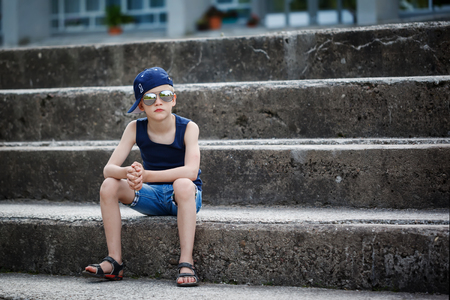 Fashionable little boy in sunglasses and cap siting on stone steps. Childhood.  Summertime.の写真素材