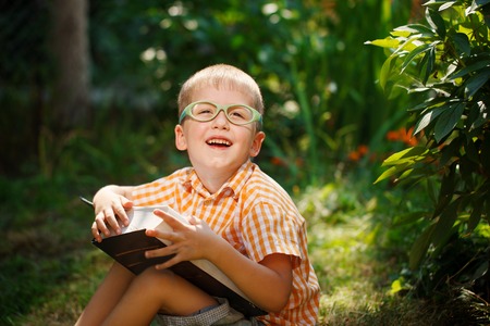 Beautiful kid boy with glasses, reading a book in garden, sitting on grass.の写真素材