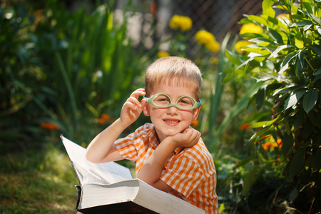 Beautiful kid boy with glasses, reading a book in garden, sitting on grass.の写真素材