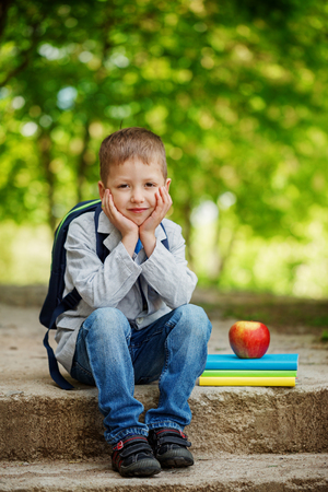 Funny  little boy sitting on stone with books, apple and backpack  on green nature background. Back to school conceptの写真素材