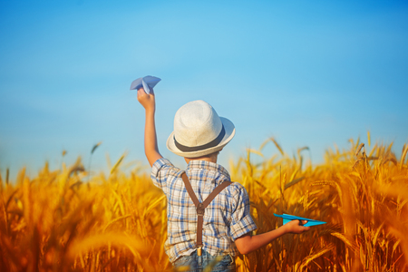 Cute child walking in the wheat golden field on a sunny summer day. 
Boy starts paper plane. Nature in the country.View from a back.の写真素材