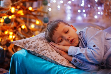 Little cute blond boy sleeping under Christmas tree and dreaming of Santa at home. Happy kid waiting for New Year and gifts.の写真素材