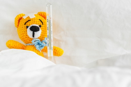 Yellow teddy bear with thermometer and plaster on head in white bedroomの写真素材