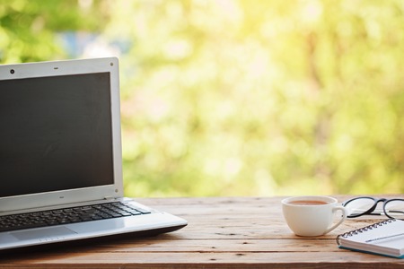 Computer with notebook, glasses and cup of tea or coffee on wooden table and nature background.の写真素材