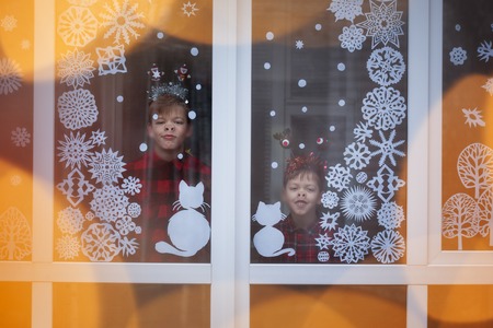 Two adorable brothers are looking through the window. They are waiting for holidays. There is decoration snowflakes from paper on the windowの写真素材