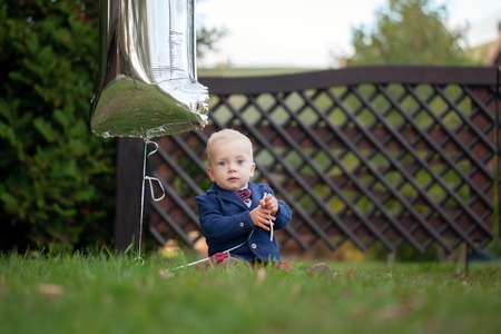 Cute playful smiled blond. 1 years old boy sitting on green grass outdor in summer dayの写真素材