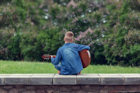 Teenager with acoustic guitar sitting on the steps in the park, outdoors. Back view.
