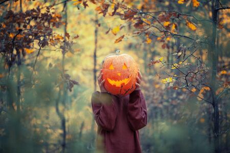 Boy holding carved pumpkin for halloween in front of his head in dark autumn forest. Halloween holiday concept.の写真素材
