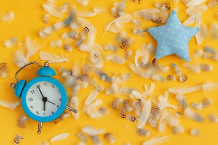 Soft blue pillow in shape of star and blue alarm clock and feathers on yellow background. Flat lay.の写真素材