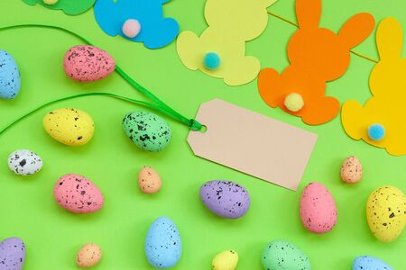 Colorful Easter eggs and handmade bunny easter garland with empty tag on green background. Top view,...