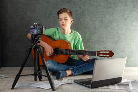 Teenager takes off the vlog of a game on a guitarat home on the camera. Young videobloger teen boy is learn to play the guitar online in living room, resting at home.の写真素材